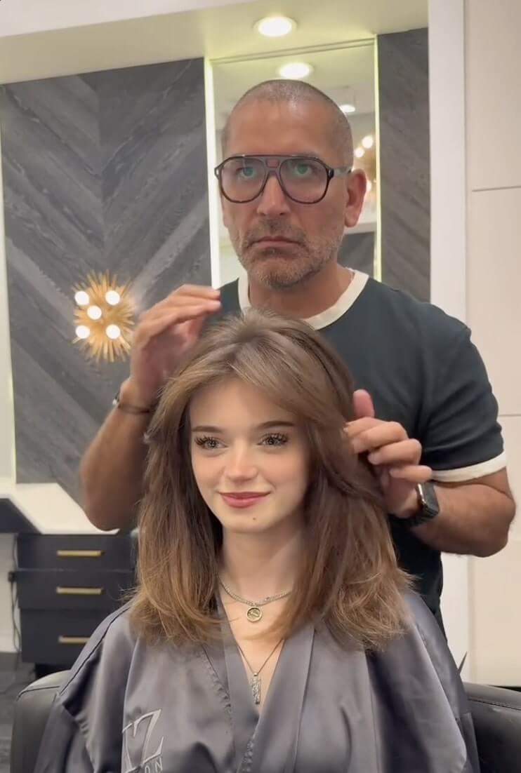 A hairstylist stands behind a seated woman with shoulder-length brown hair, adjusting her hairstyle in a modern salon.