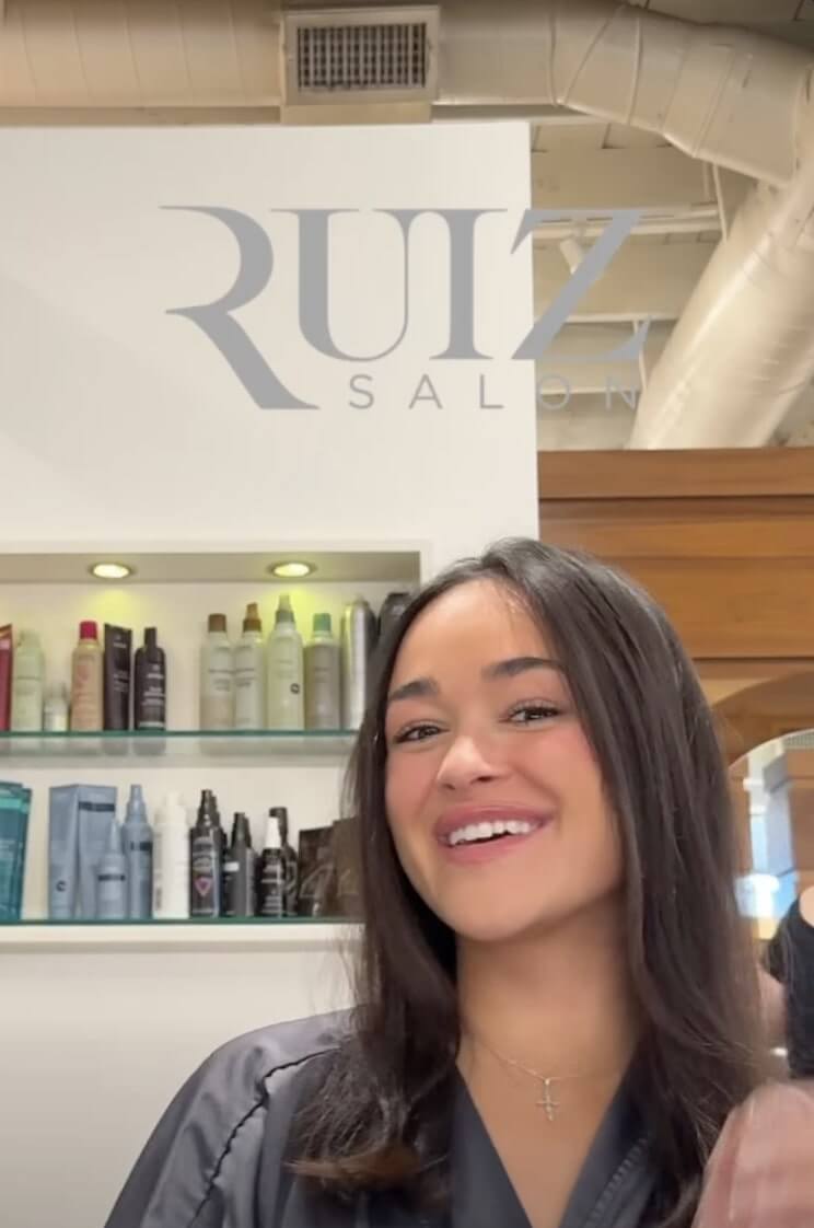 A woman with straight dark hair smiles while seated in a salon, with shelves of hair products and the "RUIZ SALON" logo visible in the background.