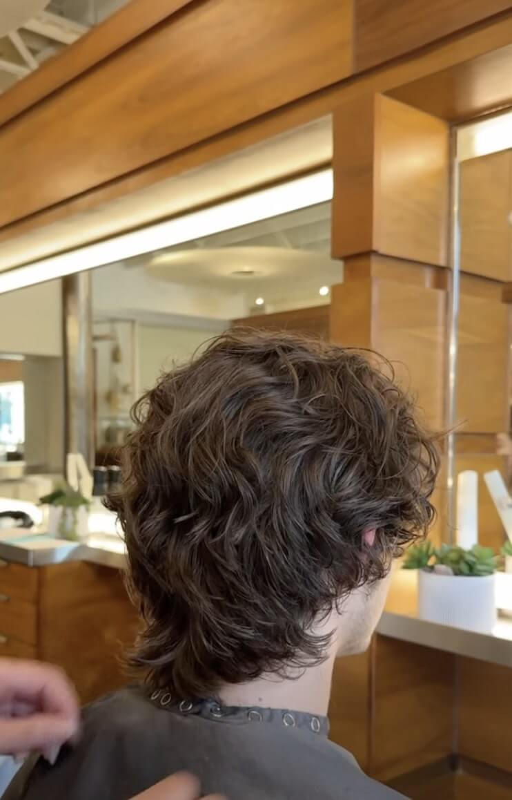 A person with medium-length, wavy brown hair sits in a salon chair, facing a mirror, as a stylist prepares to cut their hair.