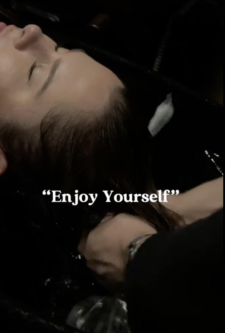 Person lying back with eyes closed receiving a hair wash, with the phrase "Enjoy Yourself" overlaid on the image.