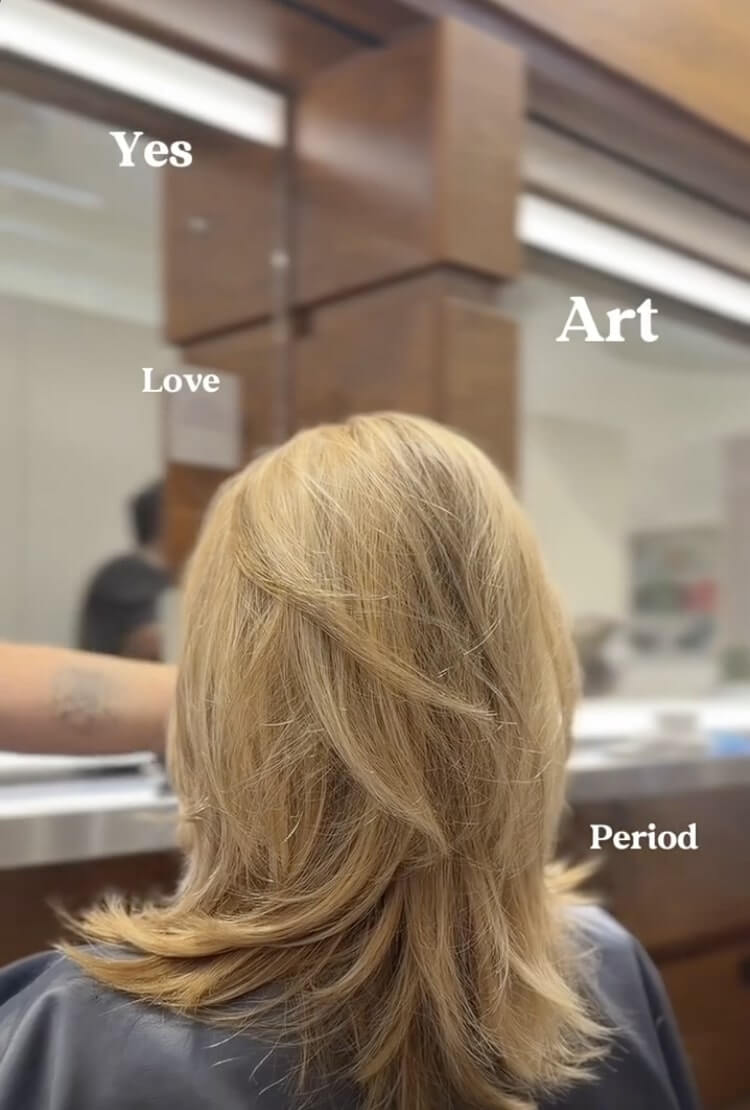 A person with shoulder-length, layered blonde hair sits facing a mirror in a salon; words "Yes," "Love," "Art," and "Period" are overlaid on the image.