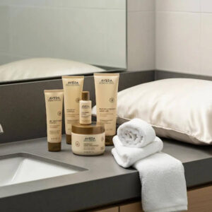 A selection of Aveda skincare products from Ruiz Salon Austin, a silk pillow, and a rolled white towel are arranged on a bathroom countertop near a sink and mirror.